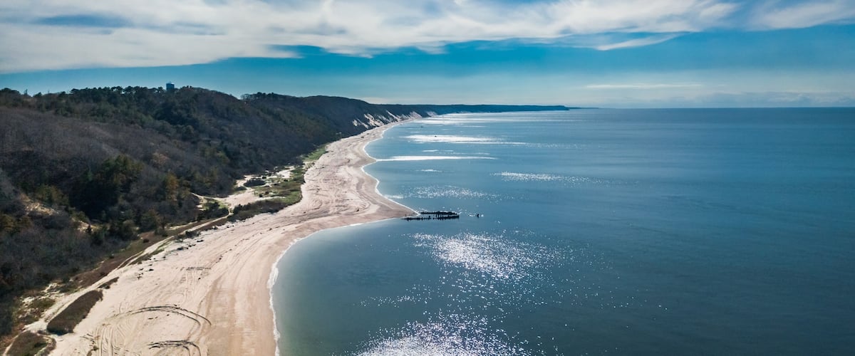 A coastal landscape with a sandy beach, a pier extending into the water, and a forested hillside.