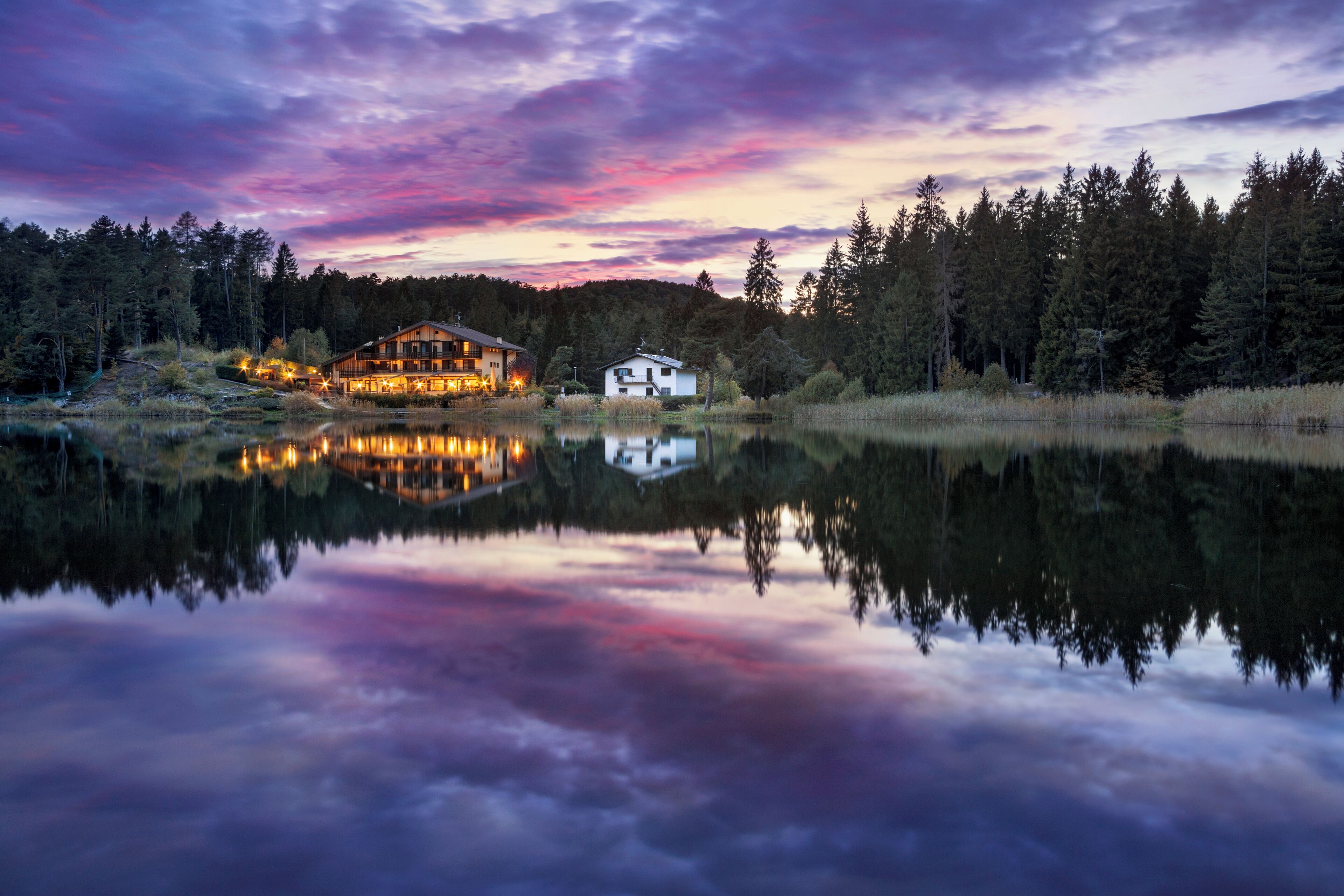 A lakeside cabin with a reflection in the water, surrounded by trees and a sunset sky.