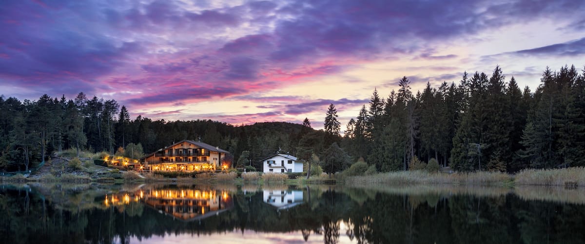 A lakeside cabin with a reflection in the water, surrounded by trees and a sunset sky.