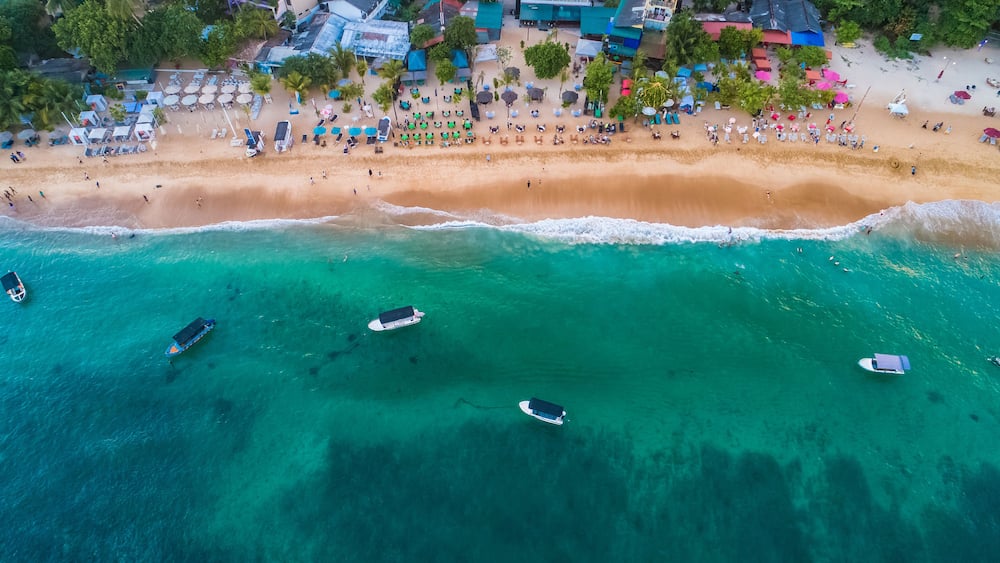 An aerial view of a beach with clear turquoise waters, several boats, and a row of beachside huts and umbrellas.