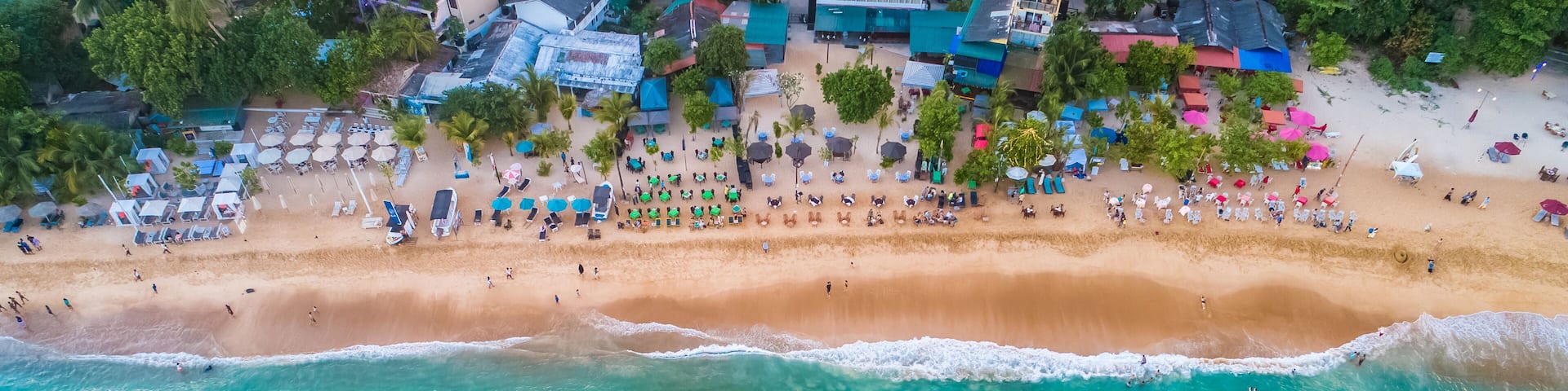 An aerial view of a beach with clear turquoise waters, several boats, and a row of beachside huts and umbrellas.