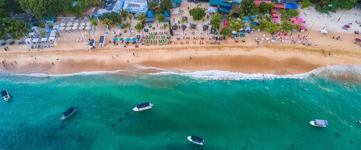 An aerial view of a beach with clear turquoise waters, several boats, and a row of beachside huts and umbrellas.
