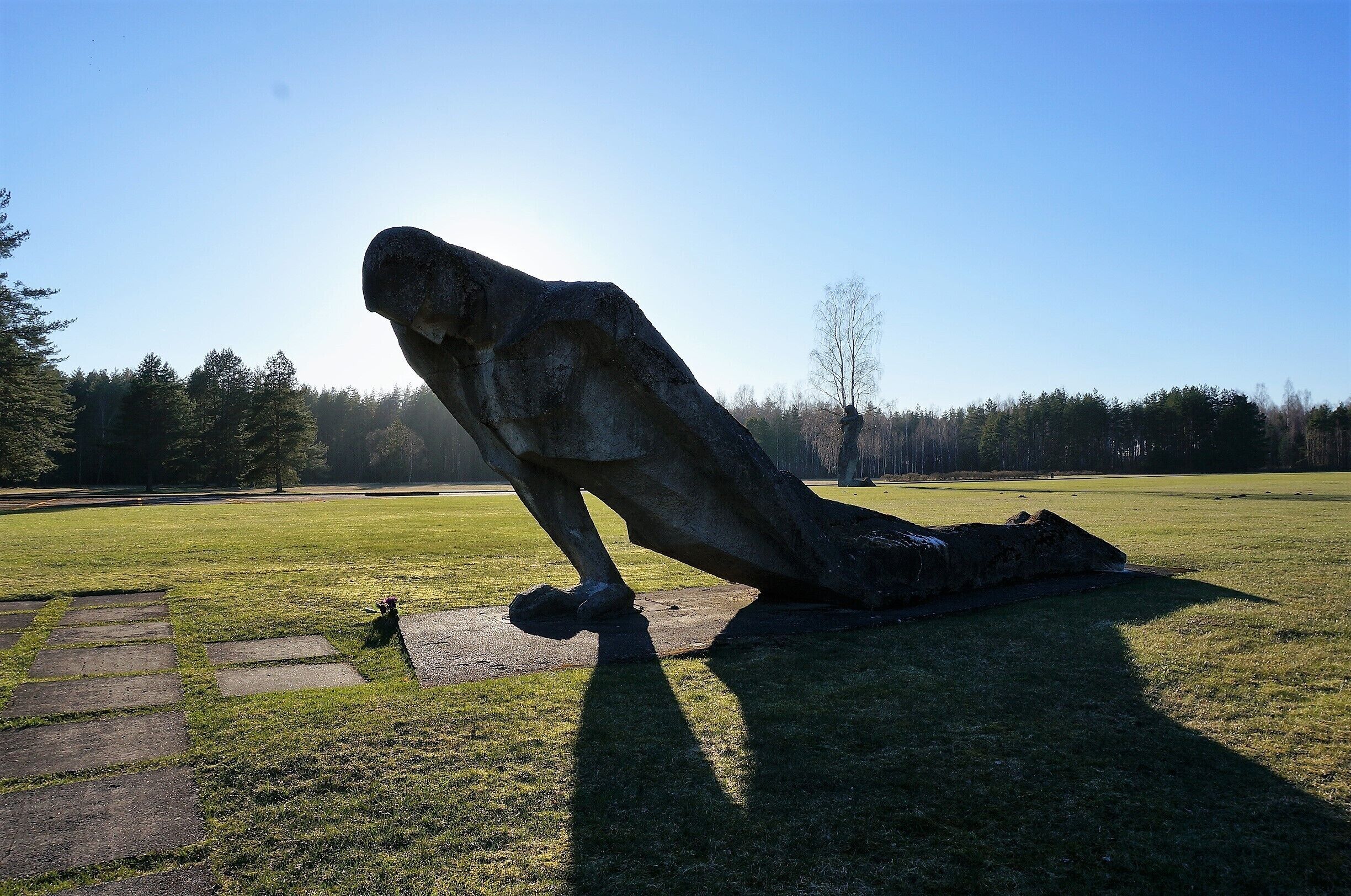 A large stone sculpture of a figure in a crouched position on a grassy field with a clear sky in the background.