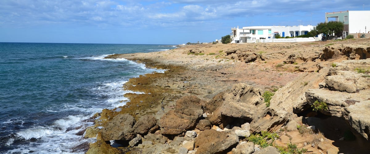 A rocky coastline with waves crashing onto the shore, a clear blue sky, and a white building with a blue door in the background.