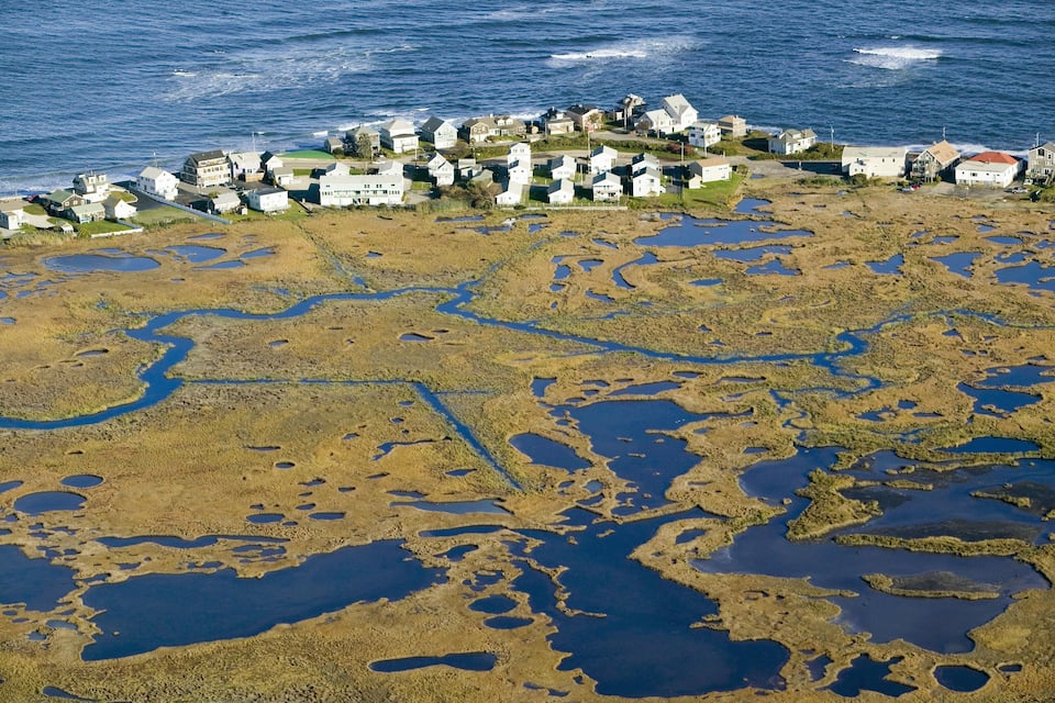An aerial view of a coastal town with houses near the water, surrounded by wetlands and tidal pools.