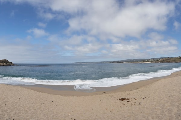 Ein Sandstrand mit sanften Wellen, einem klaren Himmel und in der Ferne Hügel.