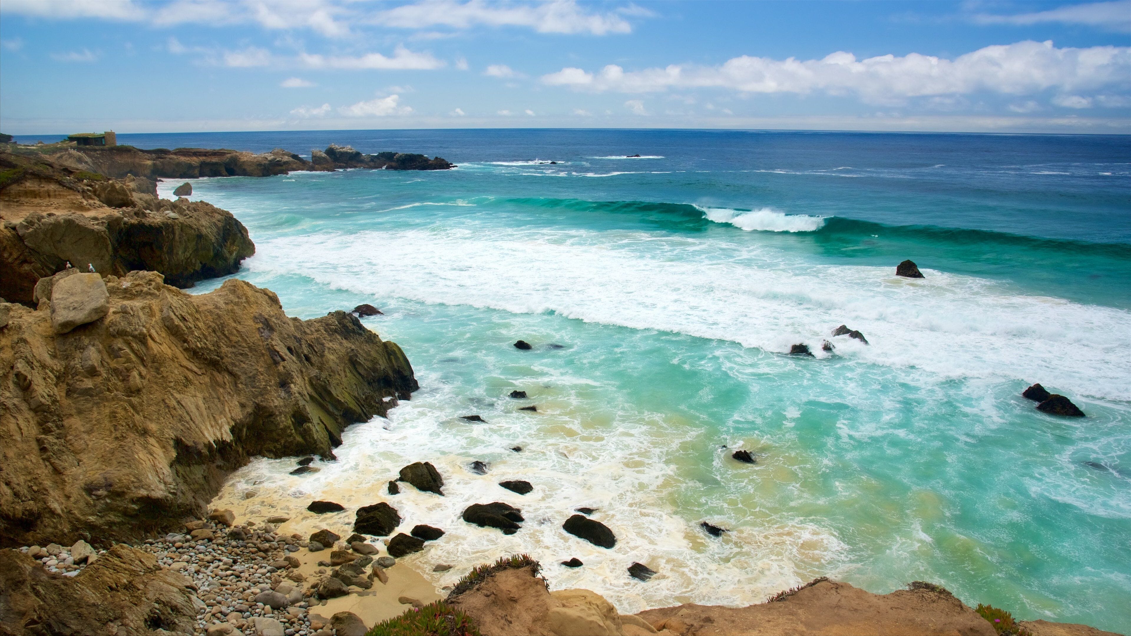 A coastal landscape with rocky cliffs, clear turquoise waters, and a distant building.