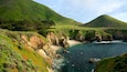 A coastal landscape with green hills, rocky cliffs, and clear blue waters.