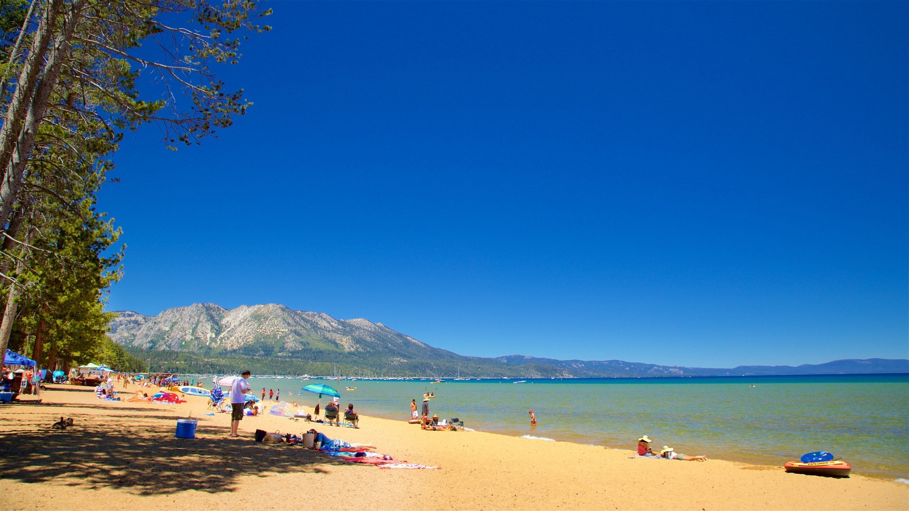 A sunny beach with people sunbathing, a boat on the water, and mountains in the background.