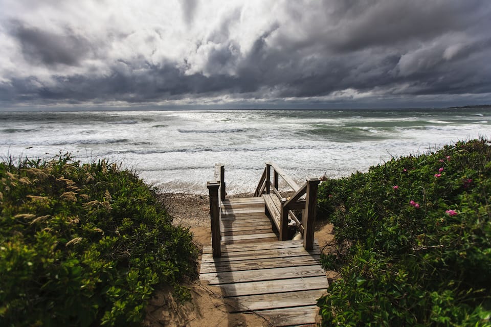 A wooden boardwalk leading to a beach with waves crashing onto the shore under a cloudy sky.