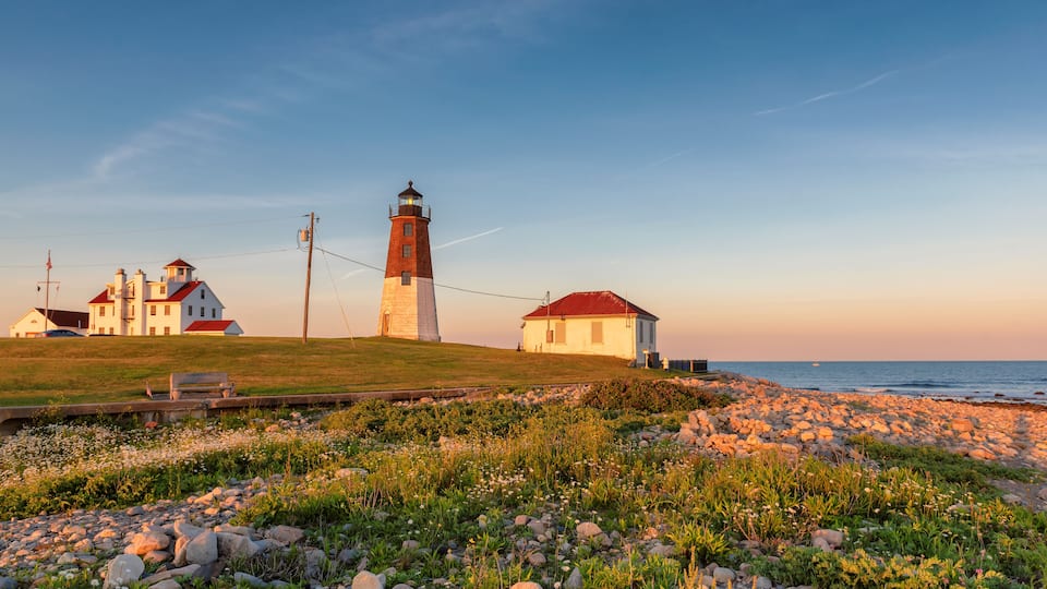A coastal landscape with a lighthouse, a white building with a red roof, and a grassy area with rocks.