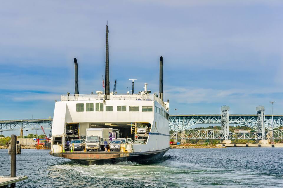 A ferry boat with multiple cars and people on board, docked at a pier with a large bridge in the background.