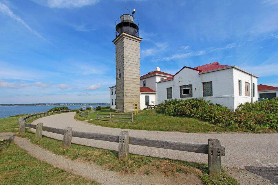 A lighthouse with a red roof, a white building with a red roof, and a body of water in the background.