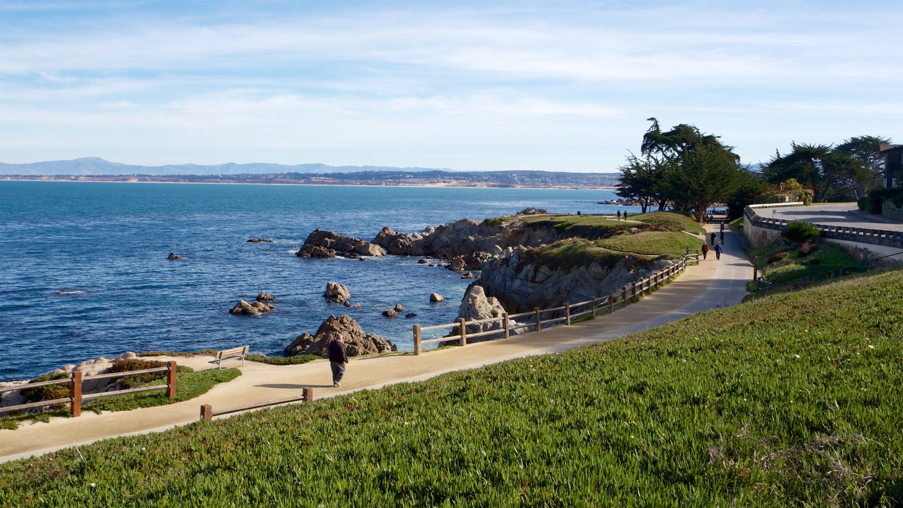 A coastal walkway with rocky outcrops, a grassy area, and a clear view of the ocean and distant mountains.
