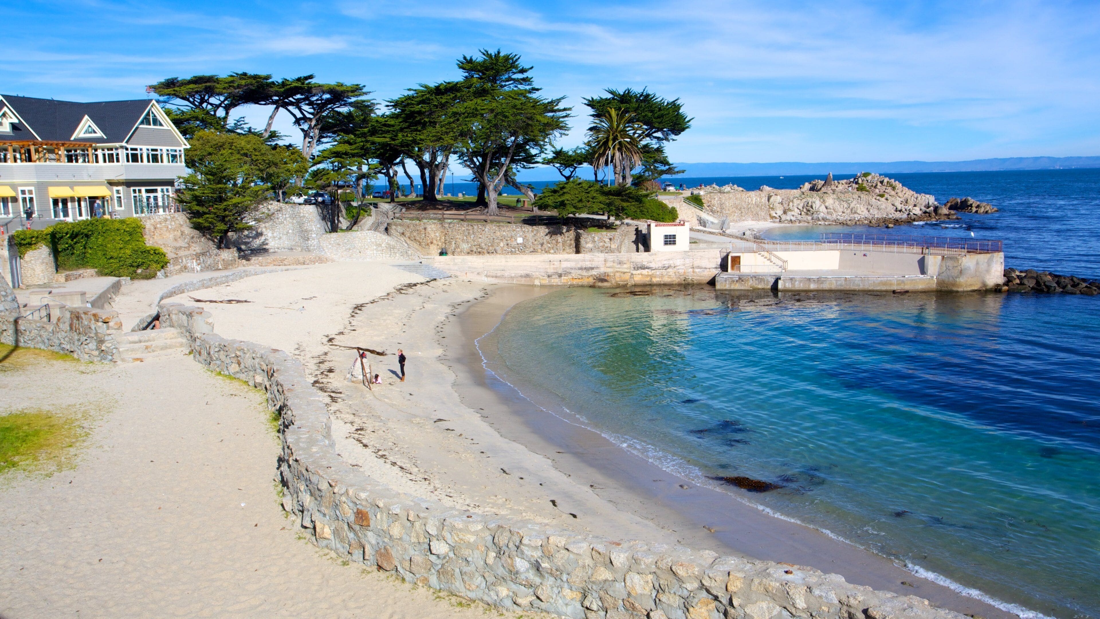 Un paysage côtier avec une plage de sable, un mur de pierre et une grande maison dotée d’un balcon donnant sur la mer.