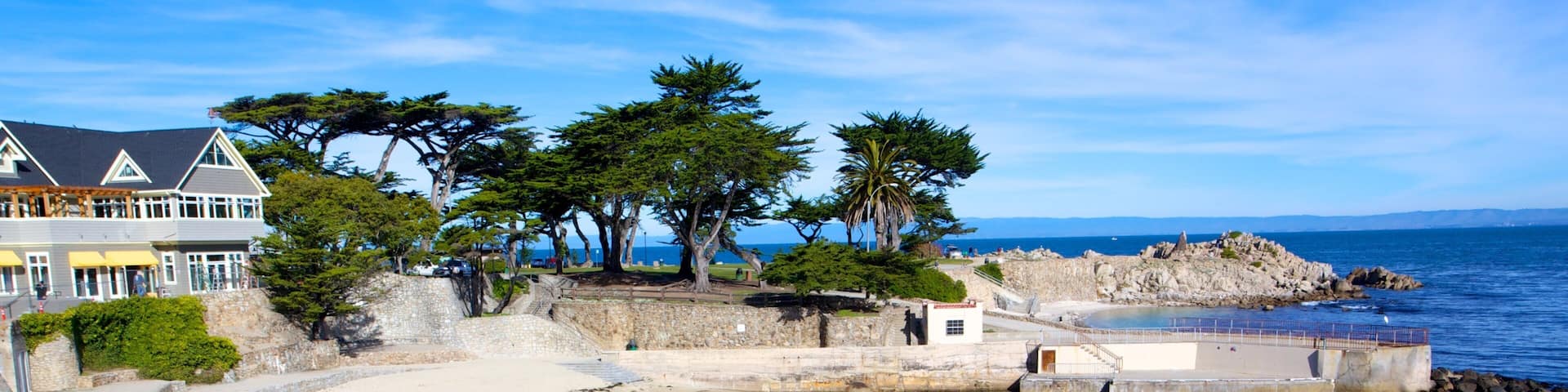 A coastal scene with a sandy beach, a stone wall, and a large house with a balcony overlooking the sea.