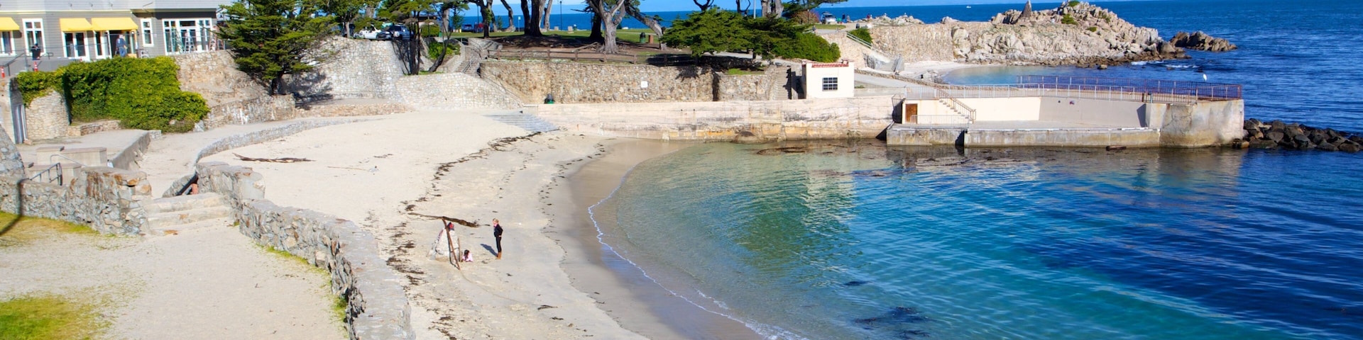 Paisaje costero con playa de arena, muro de piedra y una casa grande con balcón con vistas al mar.