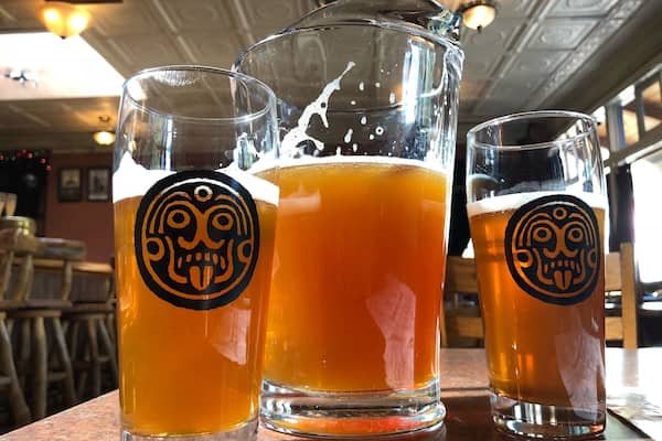 Three glasses of beer with a skull logo, one tall glass and two short glasses, on a wooden table.