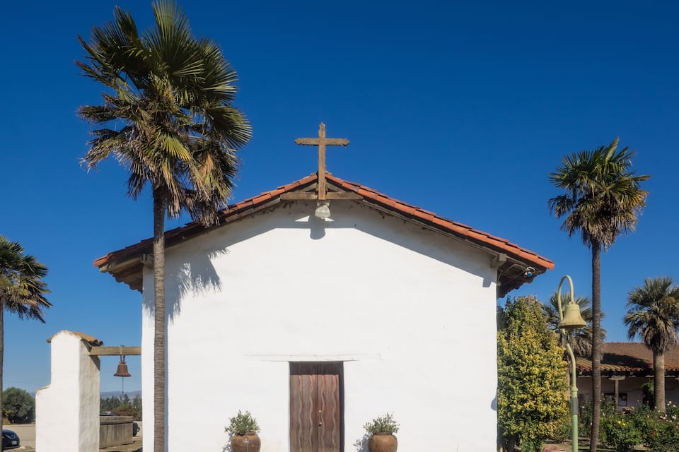 A small white church with a red roof, surrounded by palm trees and potted plants.
