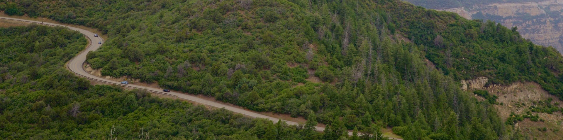 A winding road through a mountainous landscape with dense forests and distant mountains under a partly cloudy sky.