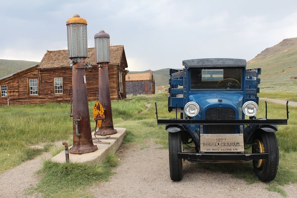 A vintage blue car parked in front of a wooden building with two rusted water towers.