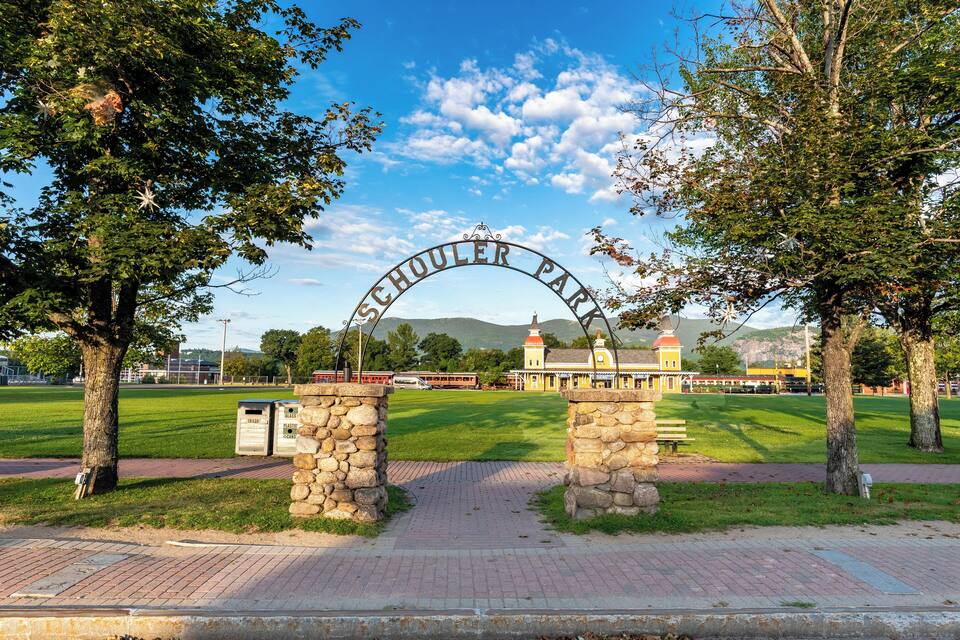 Una entrada de parque con un arco de piedra, árboles, un campo cubierto de césped y un edificio al fondo.