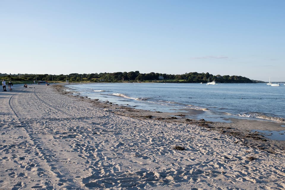 A sandy beach with gentle waves, a few boats on the water, and a forested hill in the background.