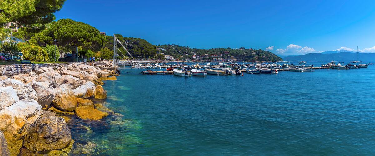 A coastal marina with boats docked, rocky shoreline, and a backdrop of hills and trees.