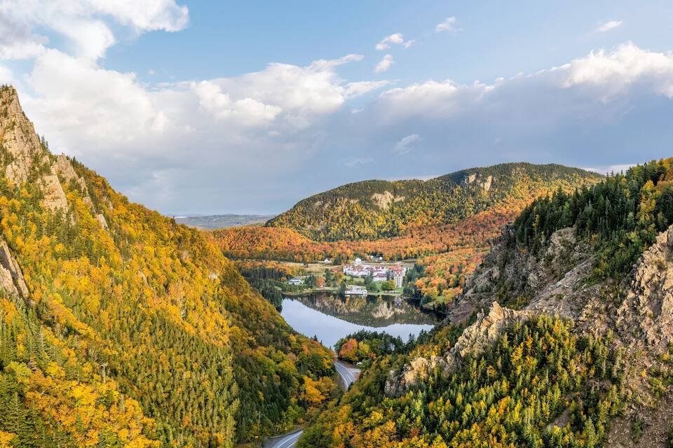Paisaje montañoso con un lago, árboles de colores otoñales y un pequeño conjunto de edificios.