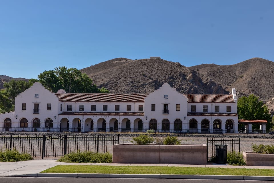 A historic white building with arched windows and a tiled roof, surrounded by a fence and set against a mountainous backdrop.