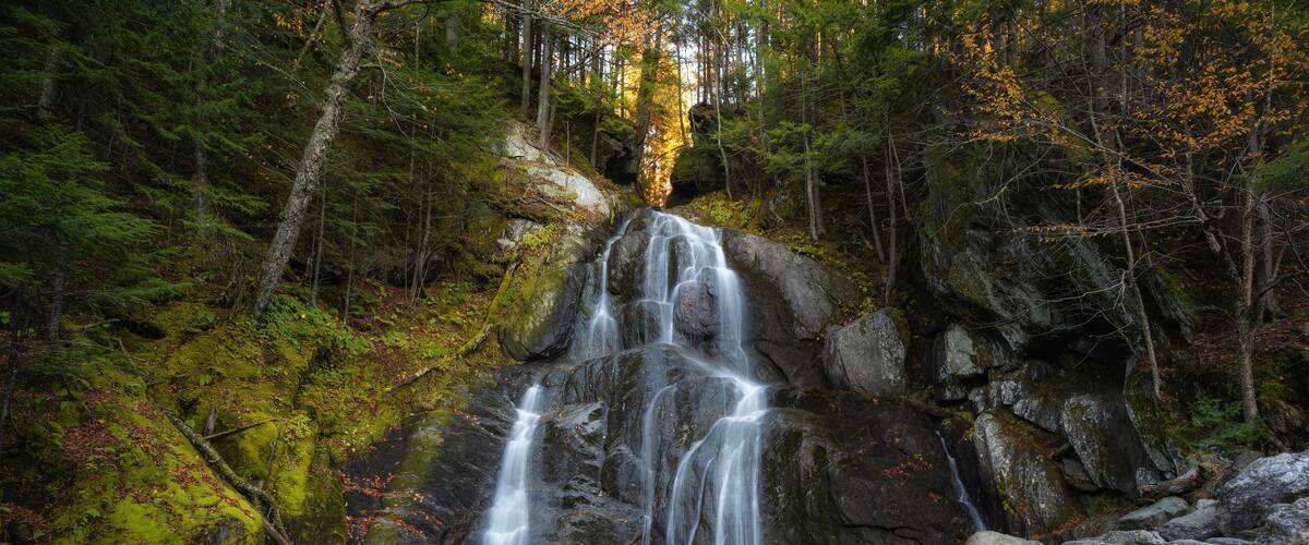 A multi-tiered waterfall surrounded by dense forest and rocky terrain.