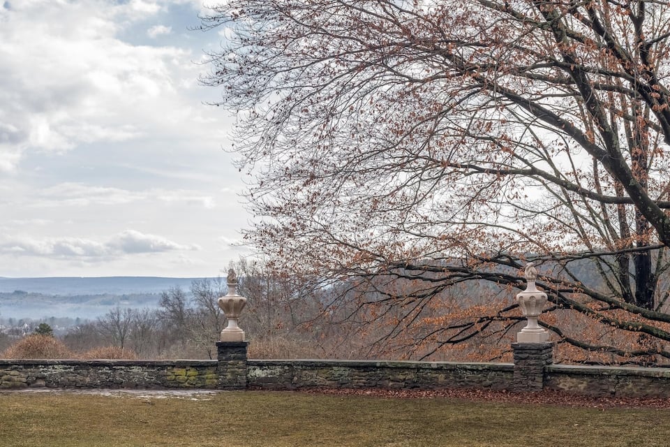 A stone bridge with ornate lamp posts, surrounded by trees and a stone wall, leading to a distant view of hills.