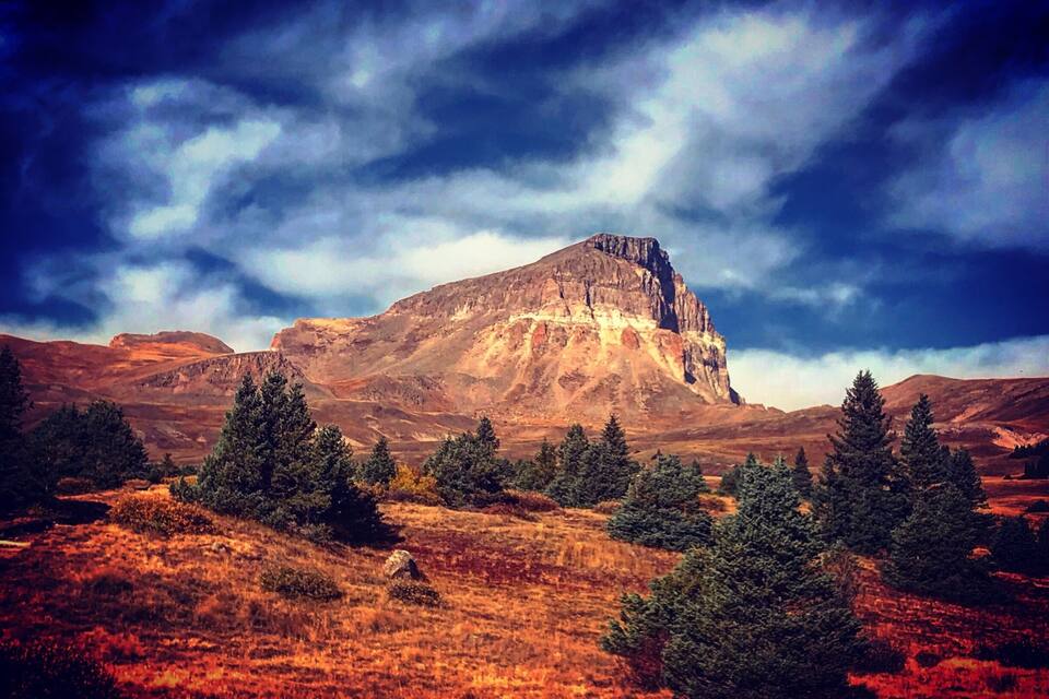 Une montagne au sommet plat, entourée d’arbres à feuilles persistantes et bordée d’une zone herbeuse au premier plan.