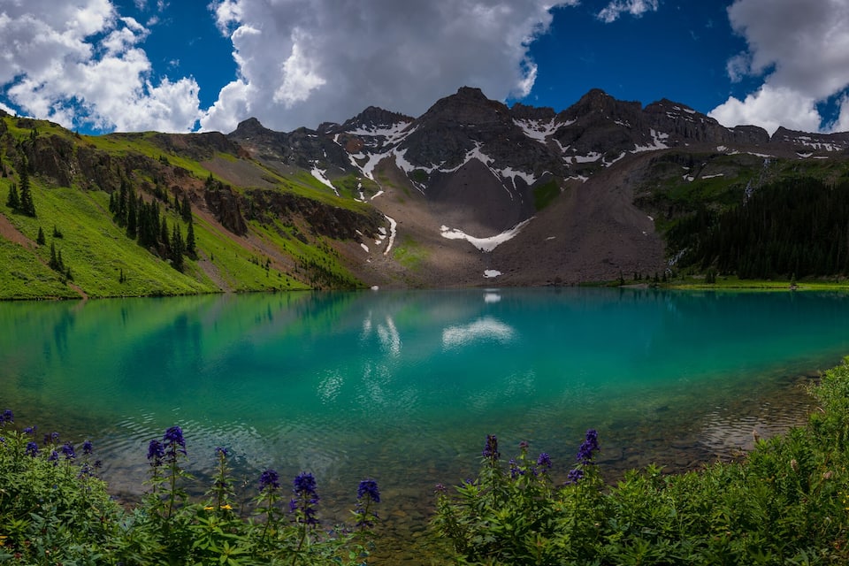 Un lago turquesa rodeado de colinas y montañas verdes con parches de nieve.