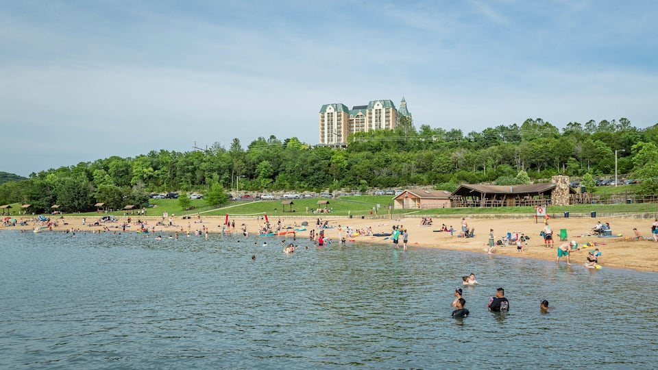 A lake with people swimming and sunbathing on the shore. There is a building with a green roof in the background.