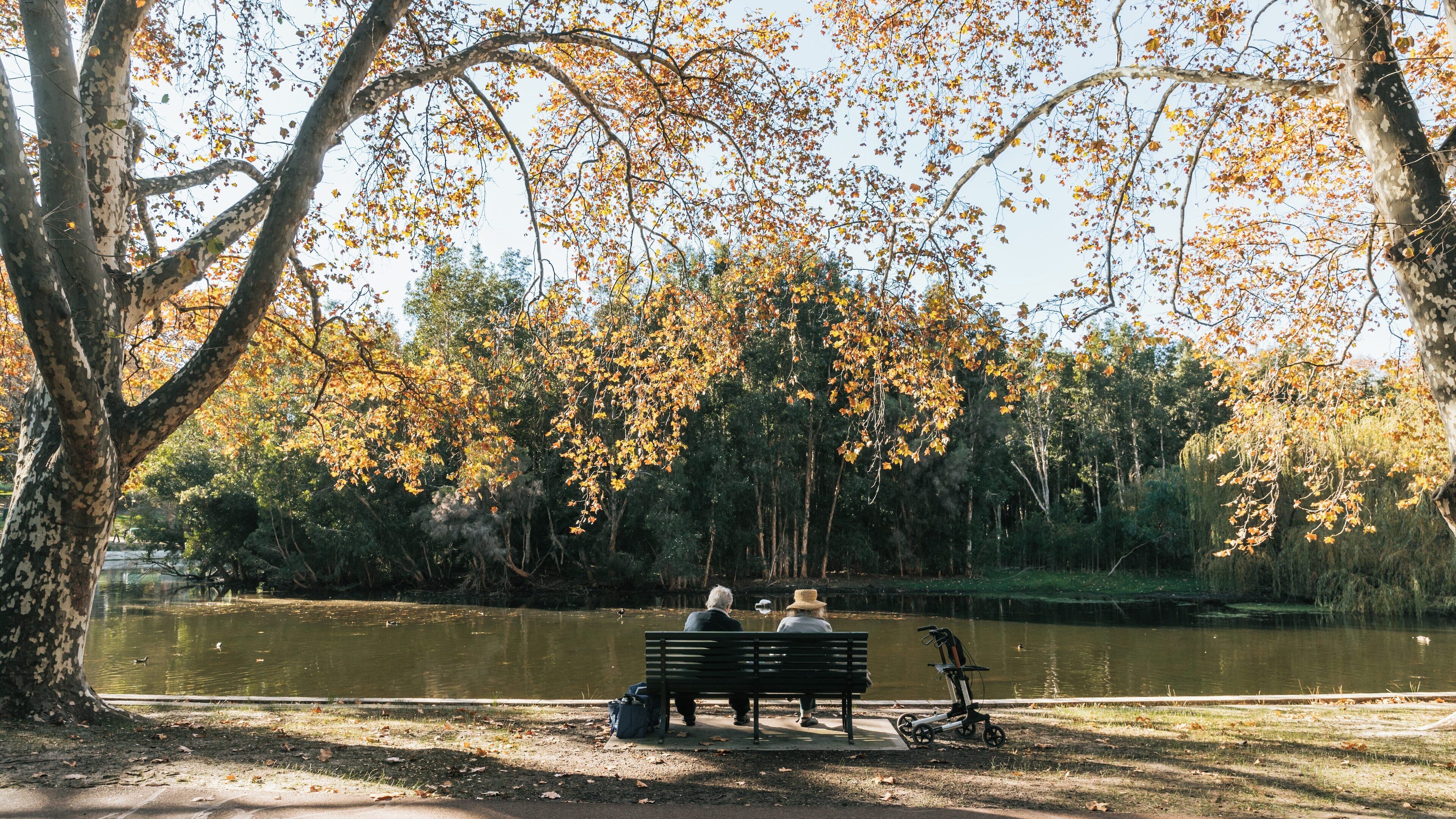 Couple seated on a bench enjoying the serene view of Hyde Park in Mount Lawley, Perth during a beautiful autumn day
