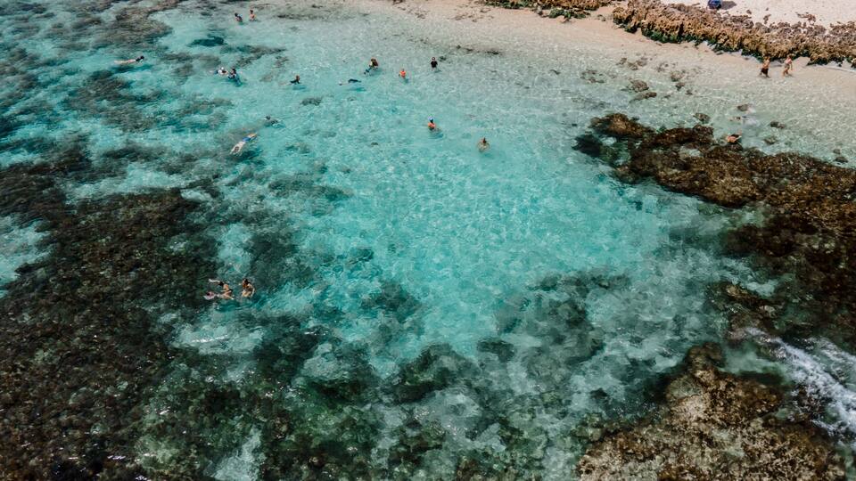 Une plage avec une eau turquoise et limpide, des formations rocheuses, et des gens qui se baignent et prennent des bains de soleil.