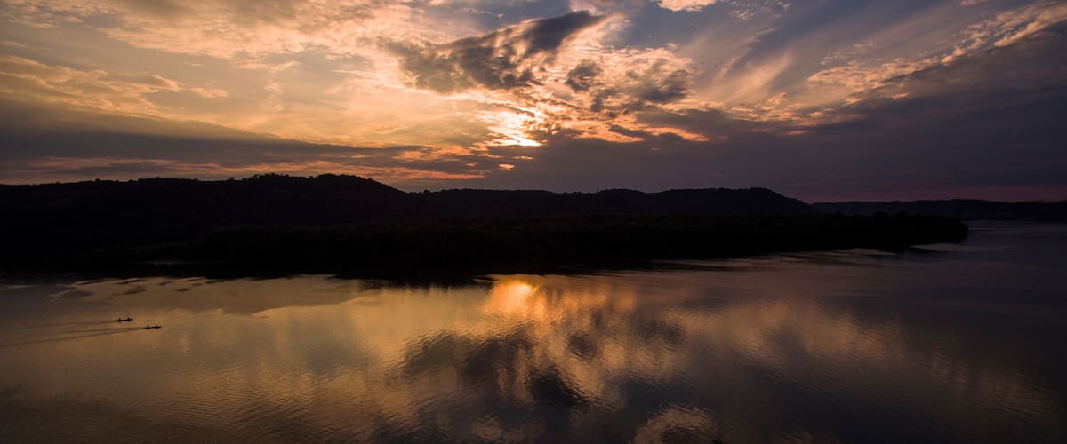 A sunset over a calm river with reflections of clouds, surrounded by hills.