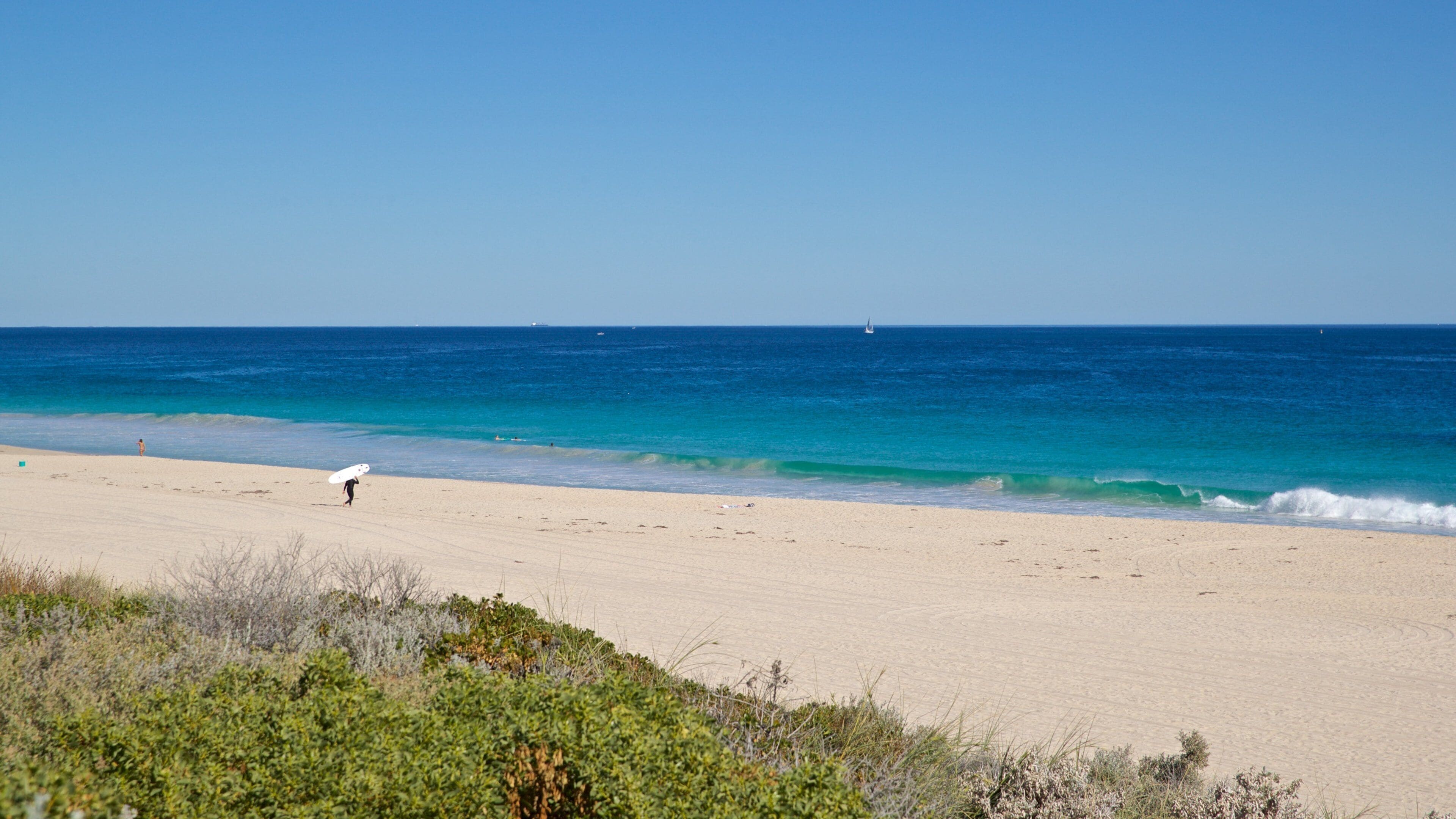 A beach with a person carrying a surfboard, clear blue water, and distant boats.