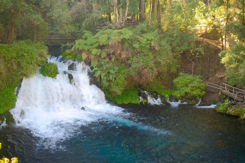 A waterfall cascading into a river surrounded by lush greenery and a wooden bridge.