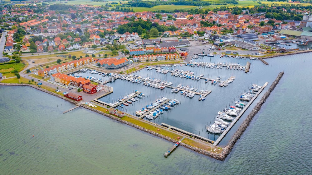 Une vue aérienne d’un port où de nombreux bateaux sont amarrés, entouré d’une ville avec des maisons aux toits rouges et un grand bâtiment au centre.