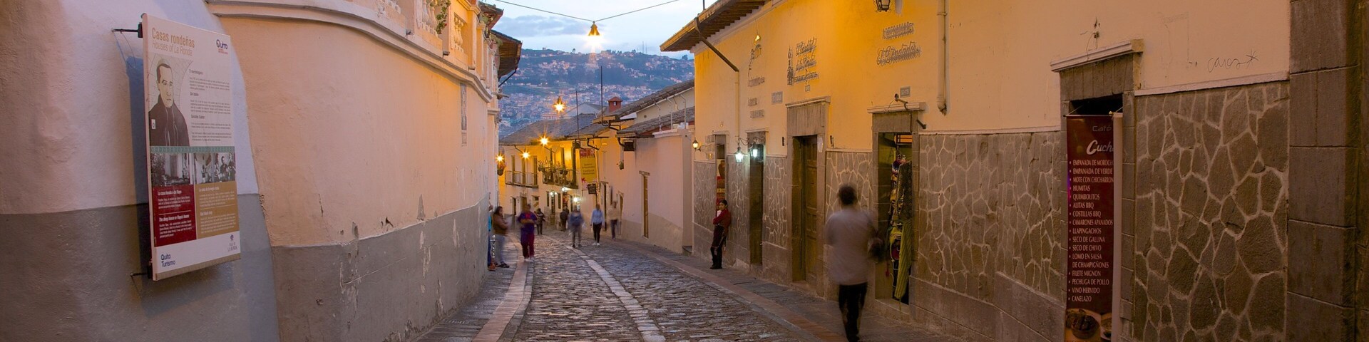 Rua de paralelepípedos ladeada por edifícios, alguns com sacadas e plantas penduradas.