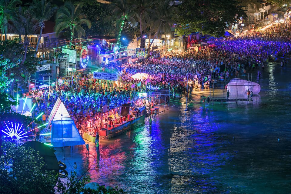 Un festival concurrido a orillas del río, con estructuras iluminadas y un bote lleno de gente.