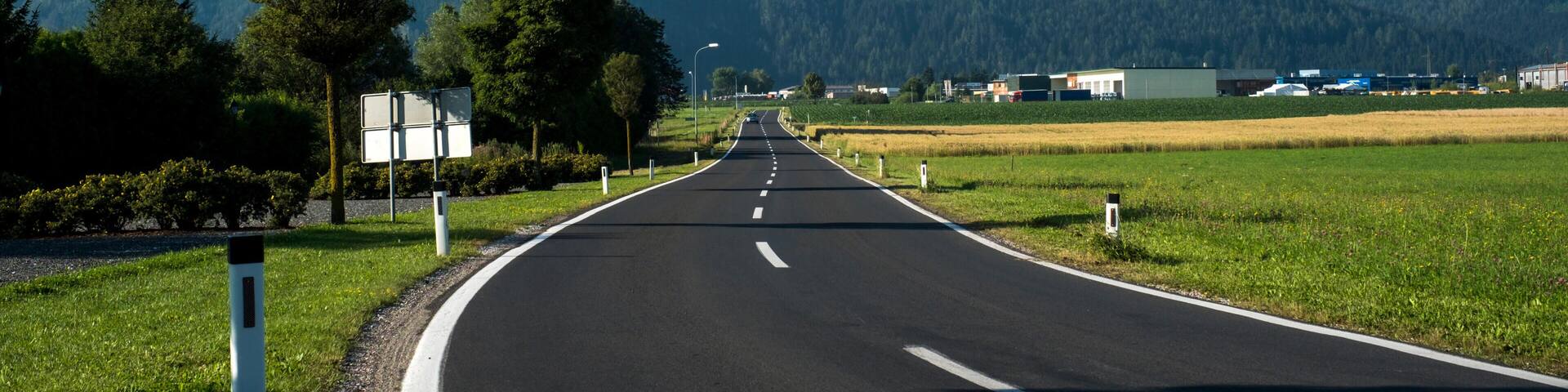 A straight road with a clear blue sky, green fields, and mountains in the distance.