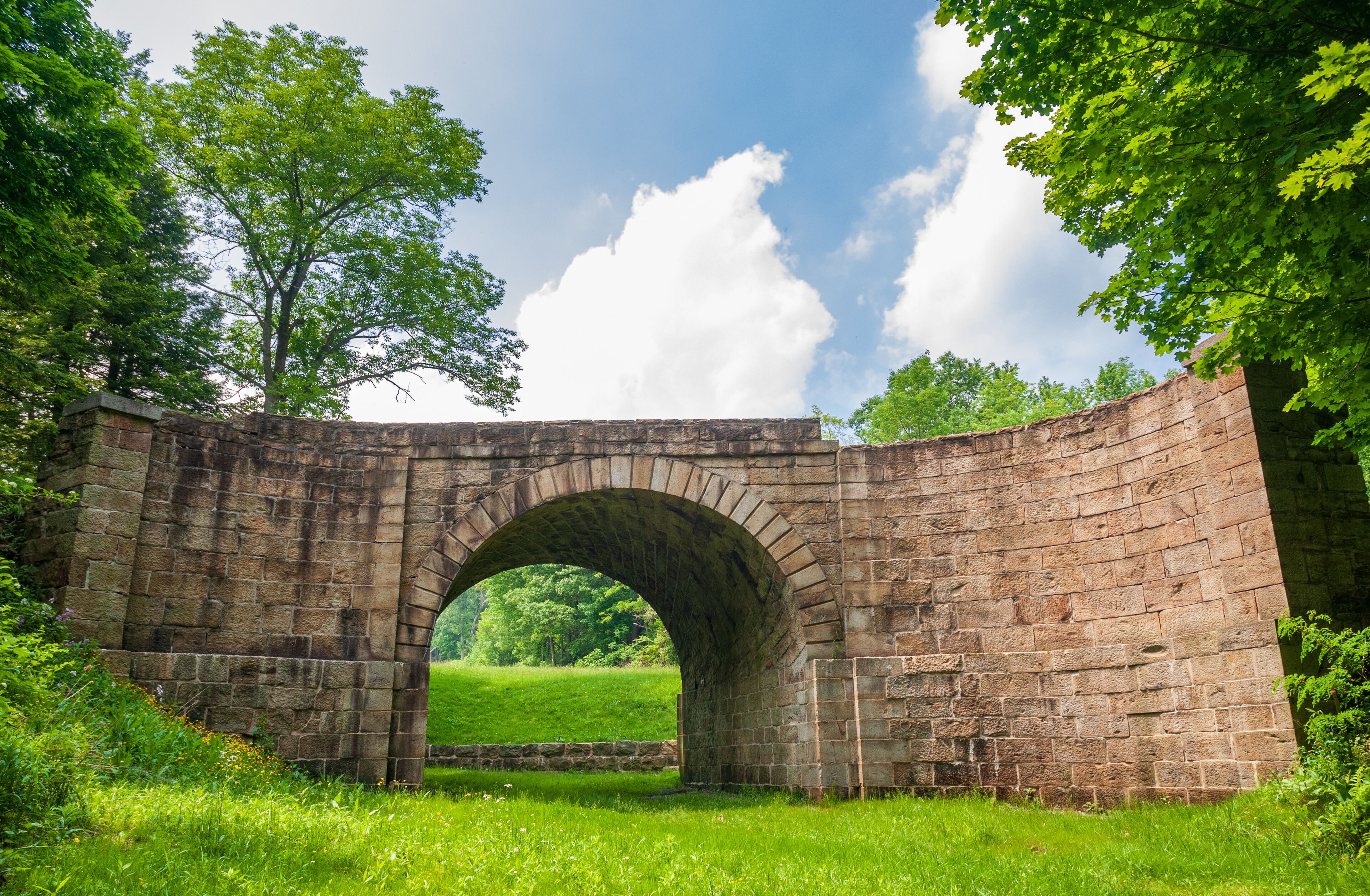An old stone bridge arching over a grassy field with trees and a clear sky in the background.
