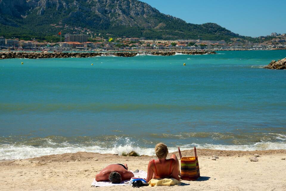 Uma praia com costa rochosa, águas turquesa calmas e montanhas ao fundo.