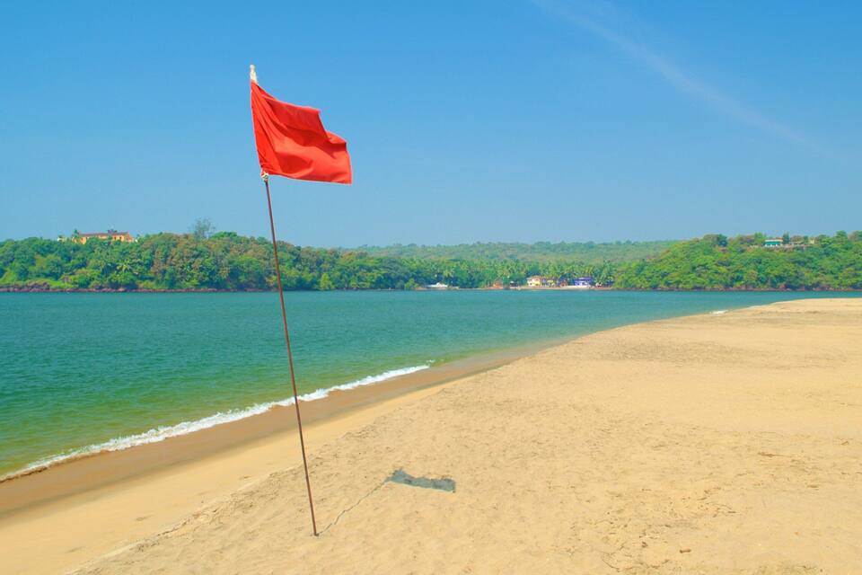 A beach with a red flag, clear blue water, and a forested hillside in the background.