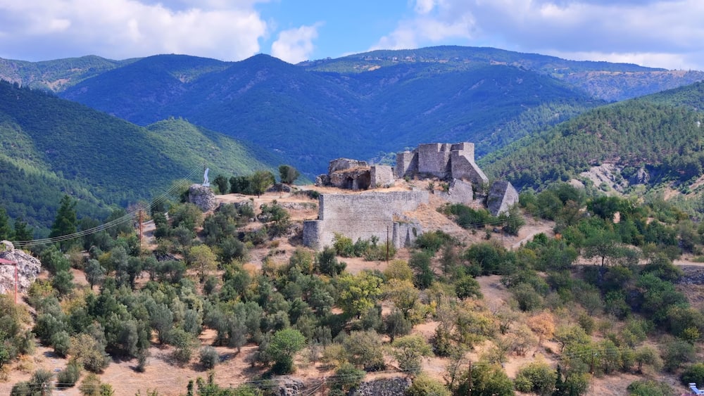 Une forteresse historique en pierre, située sur une colline et entourée d’une forêt dense et de montagnes.