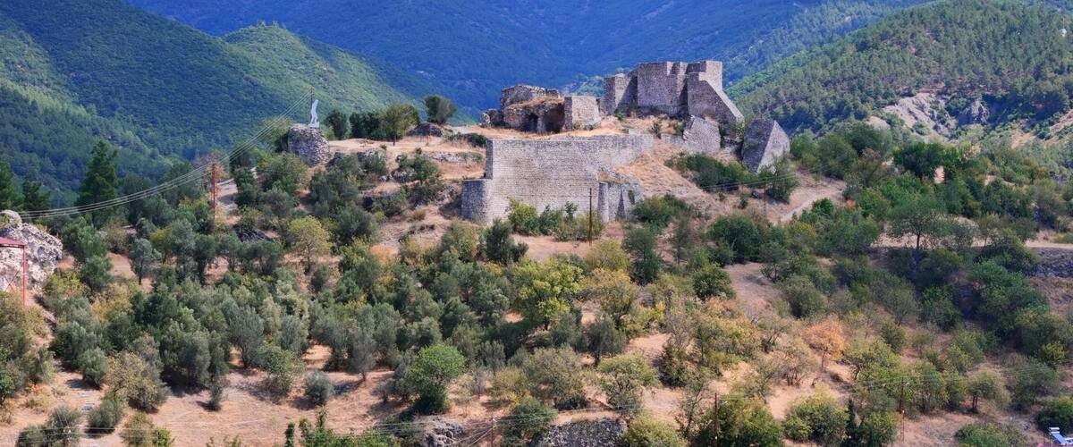 A historical stone fortress on a hill surrounded by dense forest and mountains.