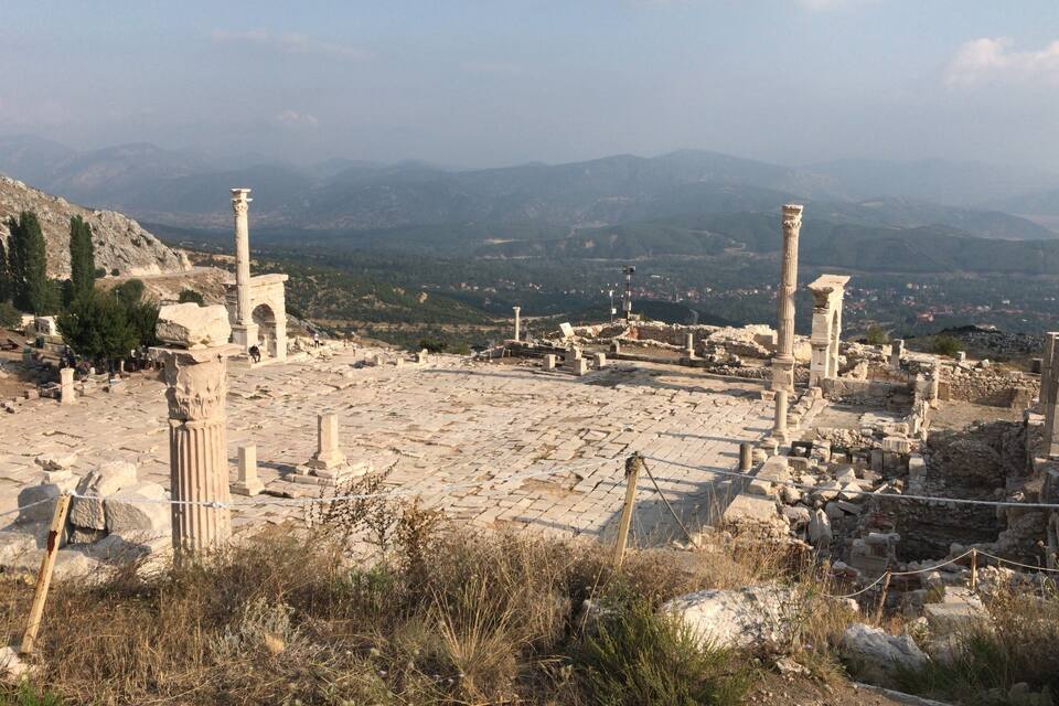 Ancient ruins with columns and stone structures, set against a mountainous backdrop.